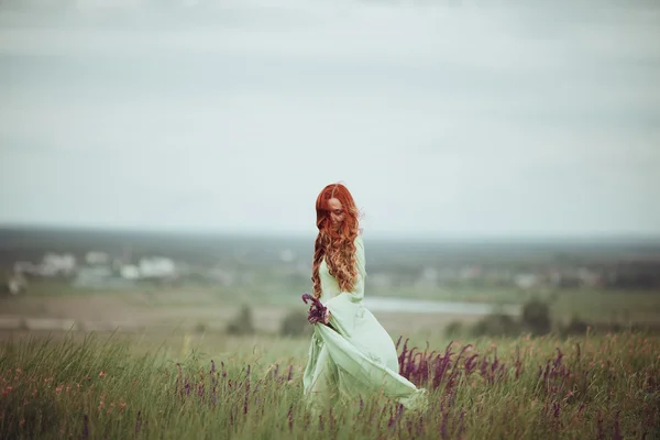 Young redhead girl in medieval dress walking through field with sage flowers. Wind concept. Fantasy