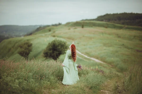 Young redhead girl in medieval dress walking through field with sage flowers. Wind concept. Fantasy