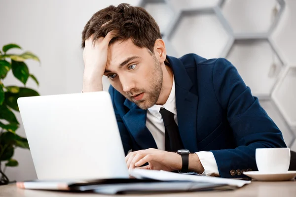 Upset young businessman sitting at laptop, office background.