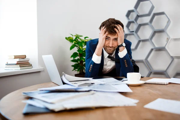 Upset young businessman sitting at workplace, office background.