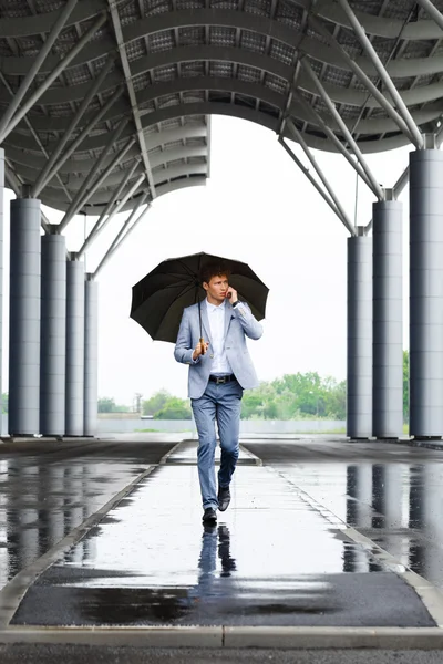 Picture of handsome young redhaired businessman holding umbrella and talking on phone walking in street