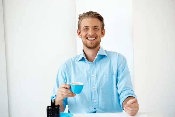 Close up portrait of young handsome cheerful smiling businessman standing at table holding coffee cup. Looking in camera. White modern office interior background.