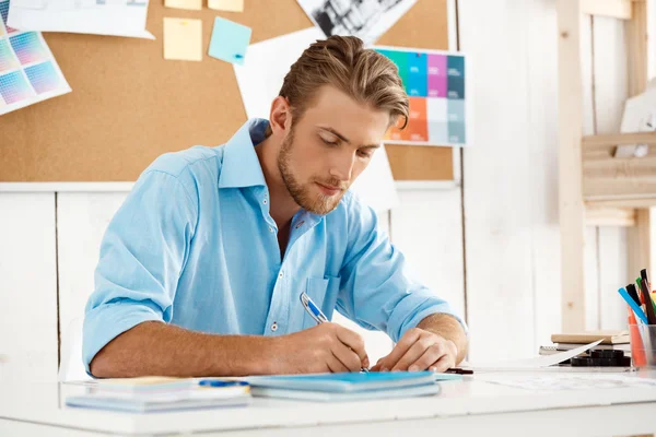 Close up portrait of young handsome confident pensive businessman working sitting at table writing in notebook. White modern office interior background.