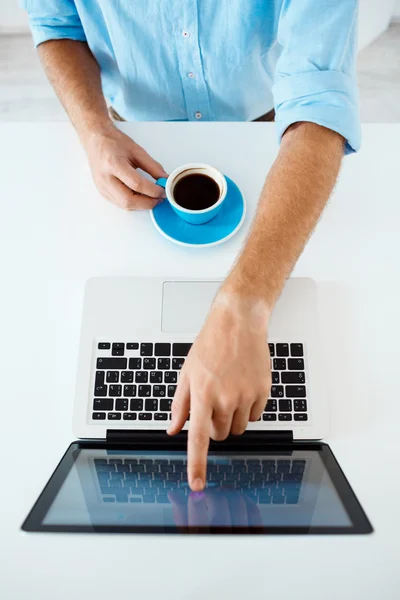 Close up picture of young businessmans hands sitting at table pointing on laptop screen holding coffee cup. White modern office interior background.