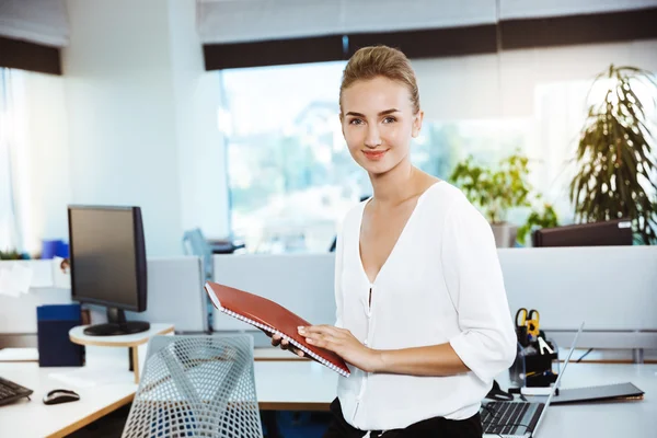 Young beautiful successful businesswoman smiling, posing, holding folder, over office background.
