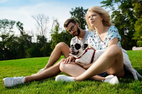 Couple smiling, sitting on grass with French bulldog in park.