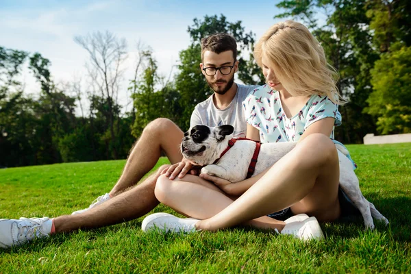 Couple smiling, sitting on grass with French bulldog in park.