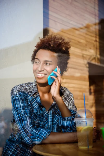 Beautiful african girl smiling, speaking on phone, sitting in cafe. Shot from outside.
