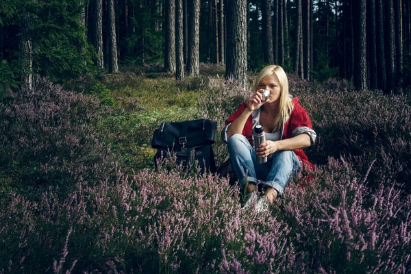 Young active girl tourist drinking tea from a thermos and sitting in the forest. Healthy active lifestyle concept