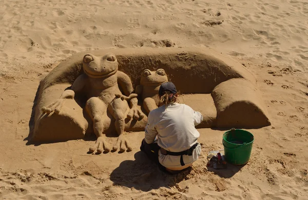 :Woman making sand sculpture of Frog aitting on sofa on Cascais beach.