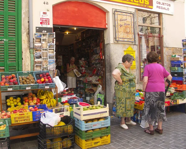 Ladies outside busy green grocers examine fruit and vegetables.