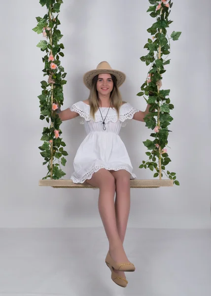 Beautiful young leggy blonde in a little white dress and white cowboy hat on a swing, wooden swing suspended from a rope hemp, rope wrapped vine and ivy