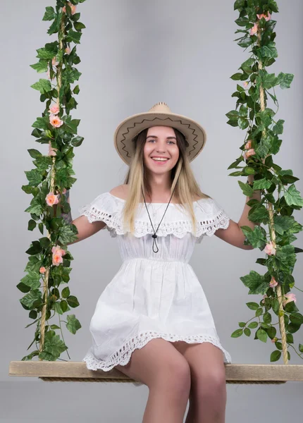 Beautiful young leggy blonde in a little white dress and white cowboy hat on a swing, wooden swing suspended from a rope hemp, rope wrapped vine and ivy