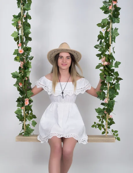 Beautiful young leggy blonde in a little white dress and white cowboy hat on a swing, wooden swing suspended from a rope hemp, rope wrapped vine and ivy