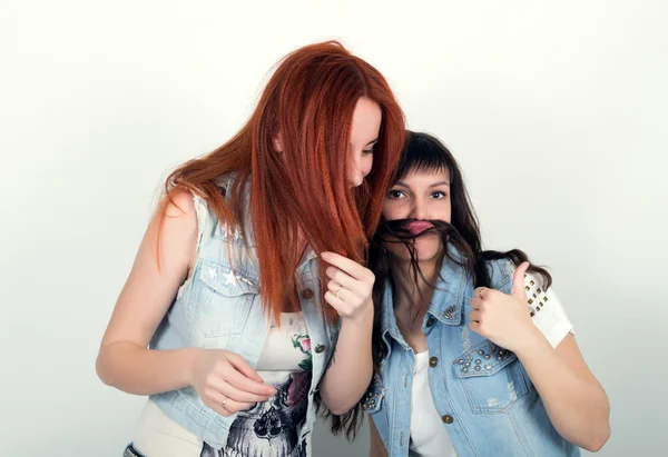 Two young girlfriends indulge and grimace, make each other a mustache out of the hair. Teenager making moustache from hair