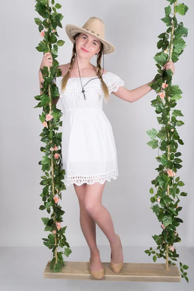 Full-length Beautiful young leggy blonde in a little white dress and white cowboy hat on a swing, wooden swing suspended from a rope hemp, rope wrapped vine and ivy