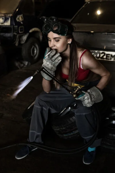 Girl mechanic sitting on a tire with blowpipe and smoking in a garage. car repairs. colorless life concept