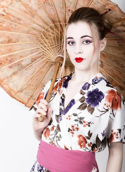 Beautiful girl dressed as a geisha girl holding a Chinese umbrella. Geisha makeup and hair dressed in a kimono. The concept of traditional Japanese values