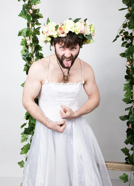 Bearded man in a womans wedding dress on her naked body, grimacing and showing tongue. on his head a wreath of flowers. funny bearded bride