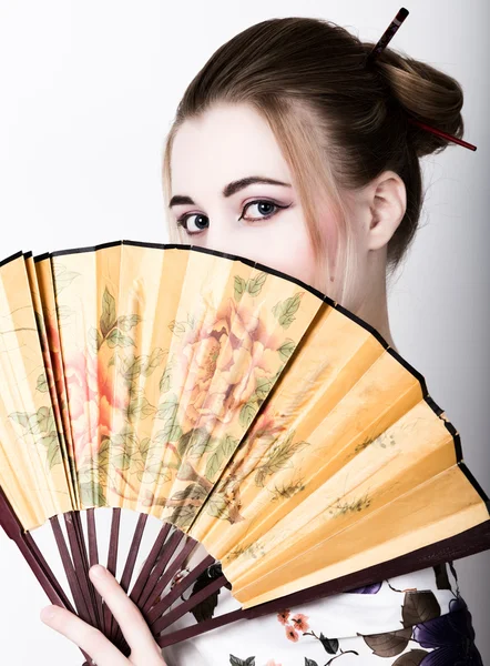 Beautiful girl dressed as a geisha, she holds a chinese fan. Geisha makeup and hair dressed in a kimono. The concept of traditional Japanese values