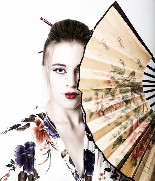 Beautiful girl dressed as a geisha, she holds a chinese fan. Geisha makeup and hair dressed in a kimono. The concept of traditional Japanese values