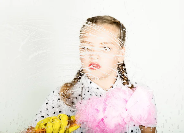 Smiling little housewife washed the window with a cloth and detergent. Large glass in foam. Housework concept.