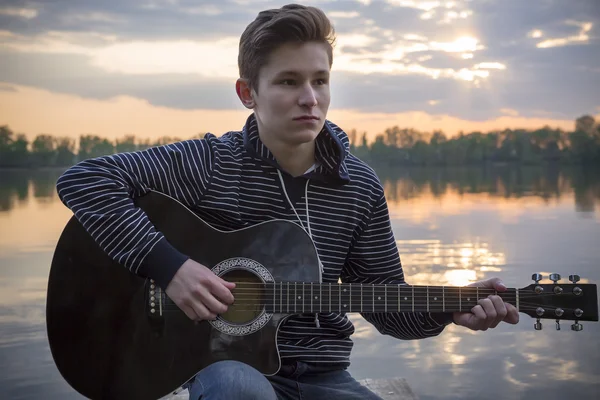 Young guy in a hood plays guitar on the bridge in the evening against the backdrop of a sunset on the river