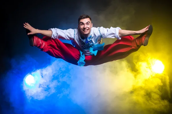 Athletic young handsome man in the Ukrainian national costume dance and jump on a dark background with smoke