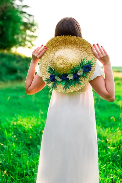 Young girl in hat with flowers stands back and looks at the sunset in the field