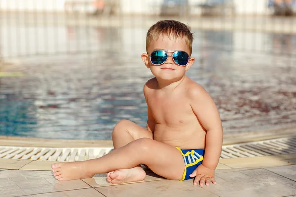 Small charming chubby baby boy in glasses sitting on a pool back