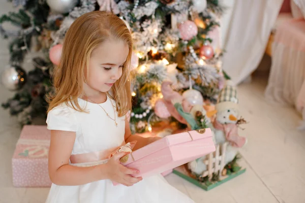 Very cute little girl blonde in a white dress holding a gift boxes on a background of Christmas trees in the interior of the house