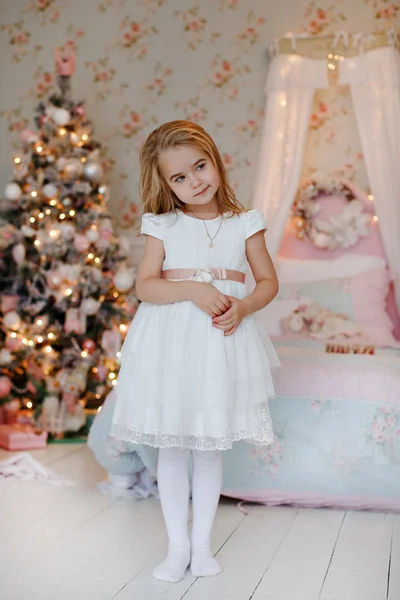 Very cute little girl blonde in a white dress holding a gift boxes on a background of Christmas trees in the interior of the house