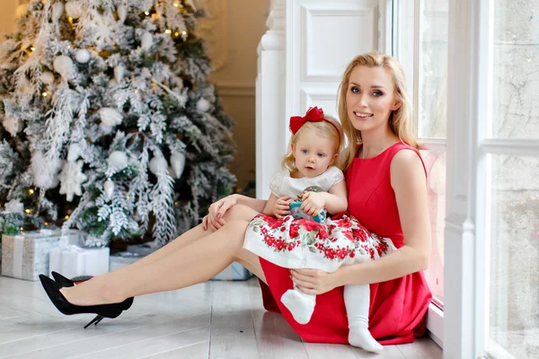 Mother in red dress smiles and holds a lovely baby girl blonde w