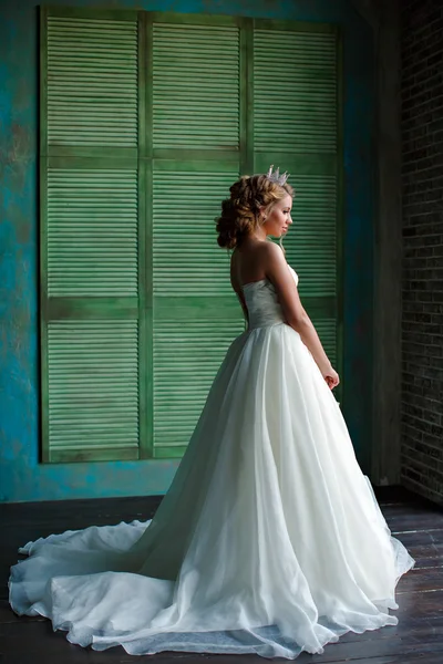 Portrait of a young blonde woman with chic thick hair, wearing a white dress and a crown on his head, looking toward the window, a full-length in profile