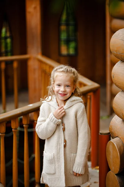 Adorable little curly blond girl in beige knitted sweater smiles  slyly, against the background of a wooden house in autumn