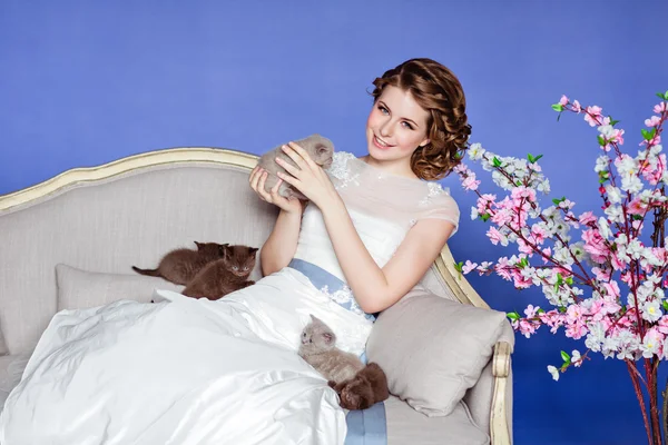 Charming and beautiful girl in white dress sitting on a sofa on a blue background with sakura flowers, smiling and holding his five little kittens