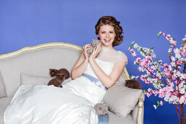 Charming and beautiful girl in white dress sitting on a sofa on a blue background with sakura flowers, smiling and holding his five little kittens