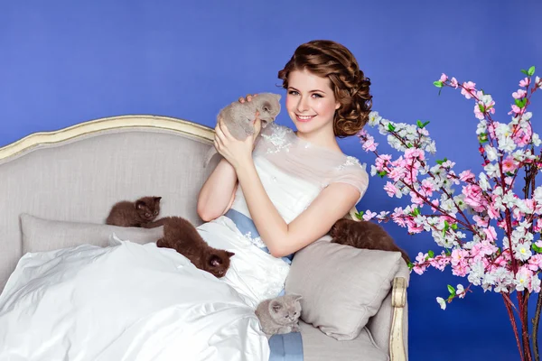Charming and beautiful girl in white dress sitting on a sofa on a blue background with sakura flowers, smiling and holding his five little kittens