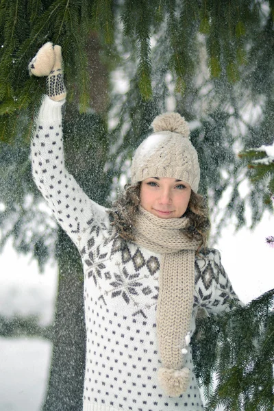 Happy,cheerful girl in winter forest,play with snow.Playful girl in snowy forest with fir-tree in warm sweater with patterns,scarf.The girl holds a fir-tree branch in a warm sweater with beautiful patterns, a cap and a scarf, shake snow and laughs.