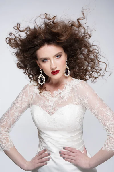 Beautiful young woman with long brown hair, red lips,jewellery in wedding dress Pretty model poses at studio.