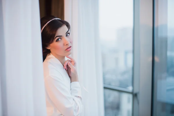 Portrait of a beautiful young girl in a white robe in the bedroom near the window