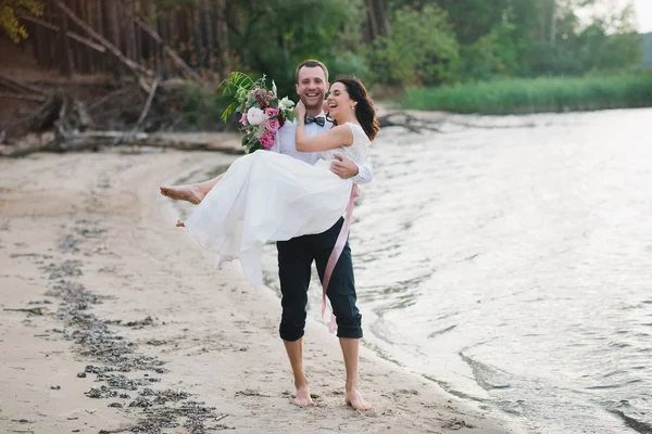 Young handsome groom holds his bride in his arms on the beach with a big bouquet of beautiful flowers