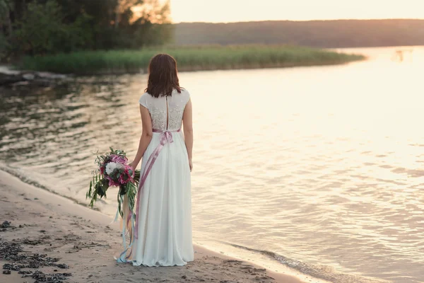 Bride standing back to nature with a bouquet in the light of sunset