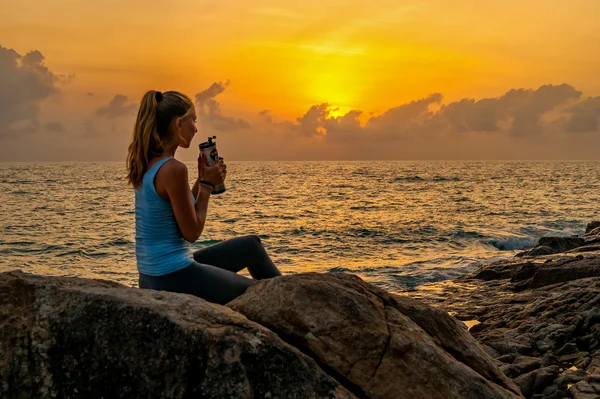 Woman sitting at dawn on the cliff near a sea,holding bottle and watching sunrise on a tropical island