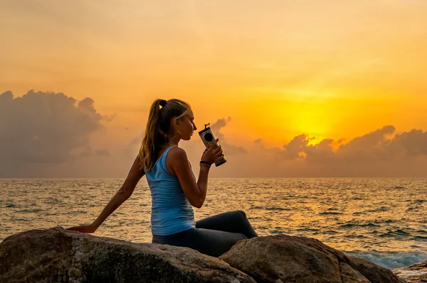 Woman sitting at dawn on the cliff near a sea,holding bottle and watching sunrise on a tropical island