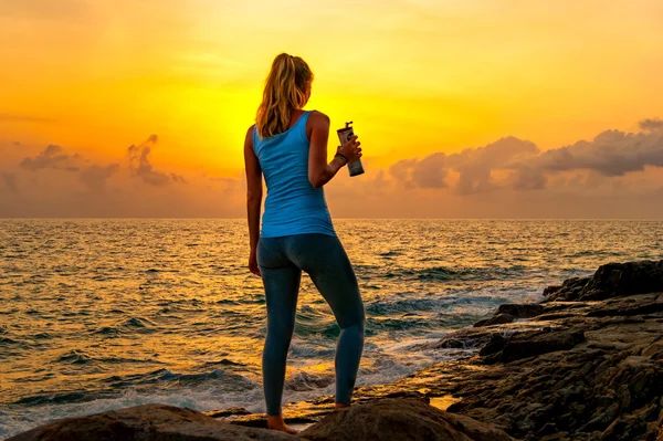 Young woman standing on the rocks by the sea, holding bottle and watching the sunrise on a tropical island