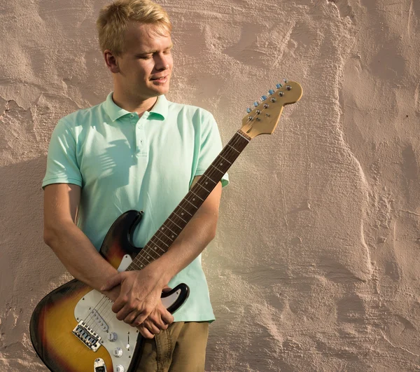 Stylish young blond man with a guitar standing against the wall, copy space