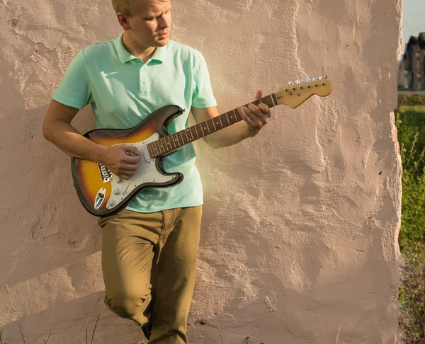 Stylish young blond man with a guitar standing against the wall, copy space
