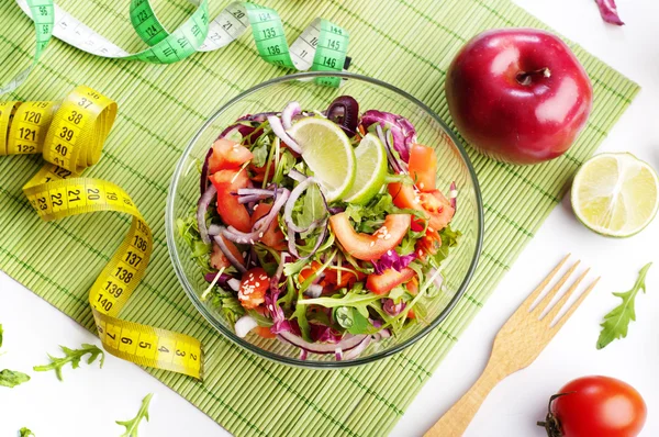 Concept diet food. Light salad of arugula, tomato, purple onion and lime on white background. Next measuring tape symbolizing measurement of body volume during a diet. Vegetarian low-fat food.