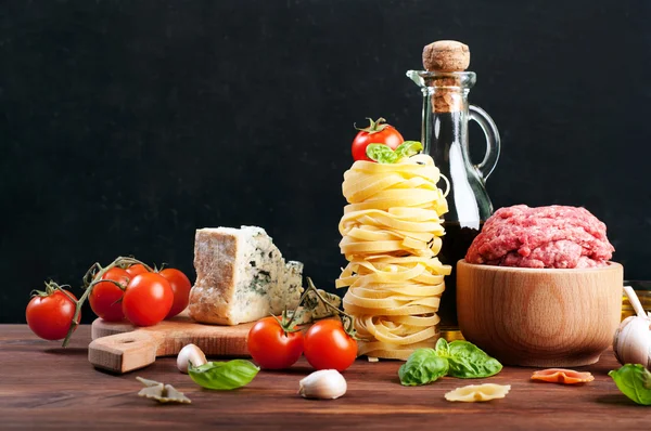 Italian food concept. Ingredients for cooking pasta, such as pasta, olive oil, ground beef, cheese and basil leaves on a brown wooden board. Black background. Place for writing text